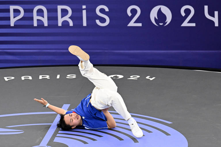 Uma atleta está realizando uma manobra de dança no chão, com uma perna levantada e o corpo inclinado para o lado. Ela veste uma camiseta azul e calças brancas. Ao fundo, há um painel com a inscrição 'PARIS 2024' em letras grandes e estilizadas.
