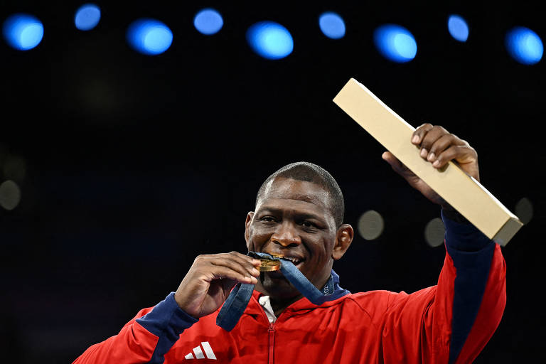 Um atleta sorridente está segurando uma medalha de ouro em uma mão e uma caixa na outra. Ele está usando um agasalho vermelho com detalhes em azul e parece estar celebrando uma vitória. O fundo é desfocado, com luzes azuis visíveis.