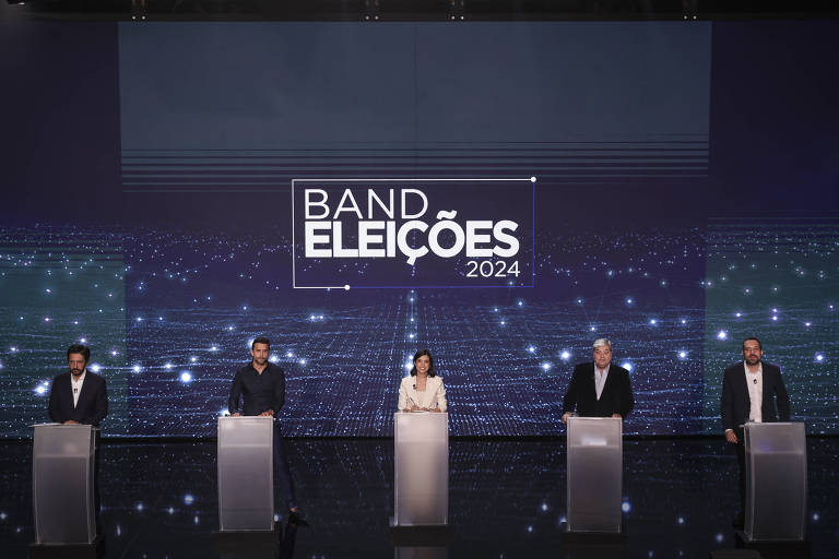 A imagem mostra um debate eleitoral com cinco candidatos em pé atrás de púlpitos. Ao fundo, há um telão com a inscrição 'BAND ELEIÇÕES 2024'. O ambiente é bem iluminado e moderno, com um design tecnológico.