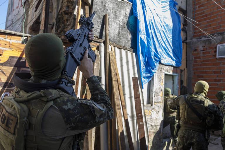 A imagem mostra soldados armados em uma operação na favela. Um dos soldados está posicionado em primeiro plano, segurando uma arma e olhando para uma construção à frente, que possui uma bandeira azul pendurada. Outros soldados estão visíveis ao fundo, se movendo em direção à mesma construção. O ambiente é caracterizado por paredes de tijolos expostos e materiais de construção.