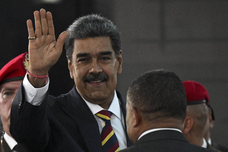 Um homem com cabelo grisalho e bigode, vestido com um terno escuro e uma gravata listrada, está acenando com a mão direita. Ele parece estar em um evento público, com pessoas ao fundo, incluindo homens com uniformes e boinas vermelhas.