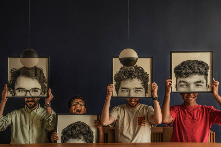 Três homens e um garoto sentam à beira de uma mesa; os dois homens mais velhos são brancos e o da ponta direita é negro; a criança é a segunda da esquerda pra direita, e segura um quadro em que aparece uma fotografia de sua testa no colo, abaixo do queixo; os outros três homens seguram quadros na frente dos seus rostos em que está fotografada do topo da cabeça até abaixo dos olhos de cada um.