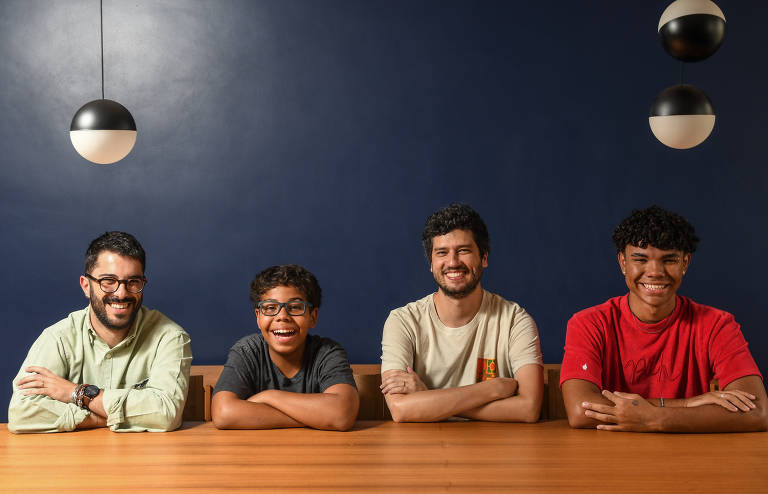 Um homem branco de óculos, um garoto negro de óculos, um homem branco e um jovem negro estão sentados à beira de uma mesa, lado a lado; eles estão com os braços cruzados apoiados sobre a mesa e todos olham para a câmera e sorriem