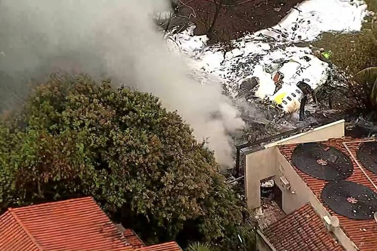 foto aérea mostra casas e, na parte do meio para cima da imagem, destroços de aeronave e coluna de fumaça. há árvores ao redor