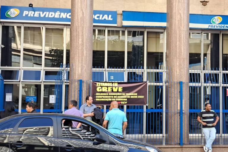 A imagem mostra a fachada de um prédio da Previdência Social, com colunas e janelas grandes. Há um cartaz em destaque que diz 'ESTAMOS EM GREVE' e menciona melhorias nas condições de trabalho. Algumas pessoas estão em frente ao prédio, algumas conversando e outras esperando. Um carro preto está estacionado na calçada.