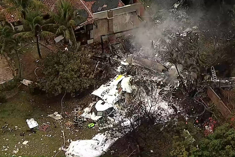 Imagem aérea de um acidente de avião, mostrando destroços espalhados em uma área verde. Há fumaça saindo dos destroços, que estão próximos a uma construção e árvores ao redor