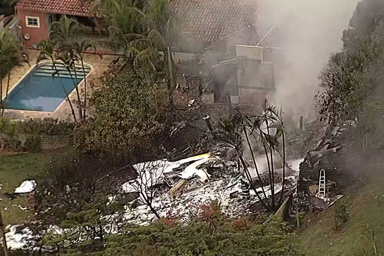 A imagem mostra os destroços de uma aeronave em chamas em uma área residencial. Há fumaça saindo dos destroços, e ao fundo, pode-se ver uma piscina e algumas árvores ao redor. A cena sugere um acidente aéreo que ocorreu próximo a uma casa.