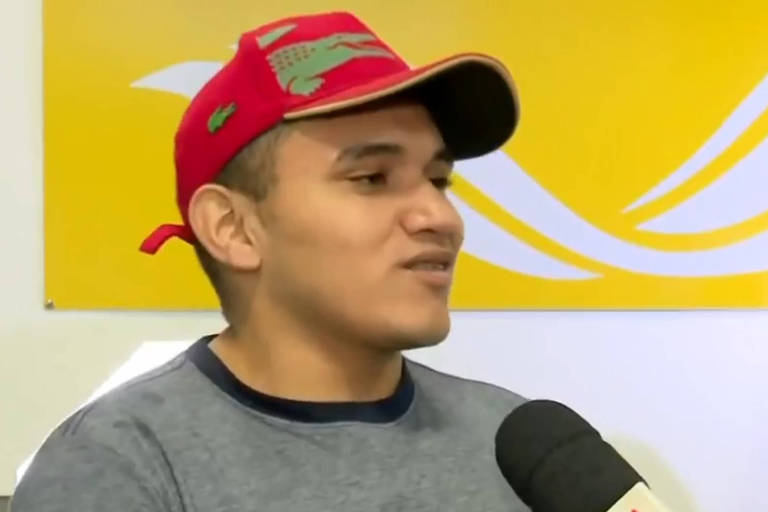 homem boné vermelho com detalhes em verde, camiseta cinza com gola preta, dá entrevista falando ao microfone, atrás dele há um painel amarelo com detalhes em branco 