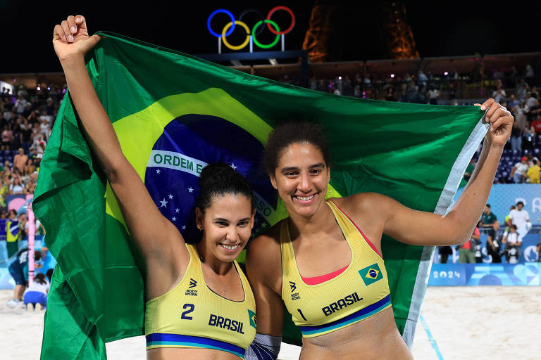 Duas atletas brasileiras estão celebrando, segurando a bandeira do Brasil. Elas estão vestindo tops amarelos com detalhes verdes e azuis, representando a seleção brasileira. Ao fundo, há uma multidão e os anéis olímpicos visíveis no céu.
