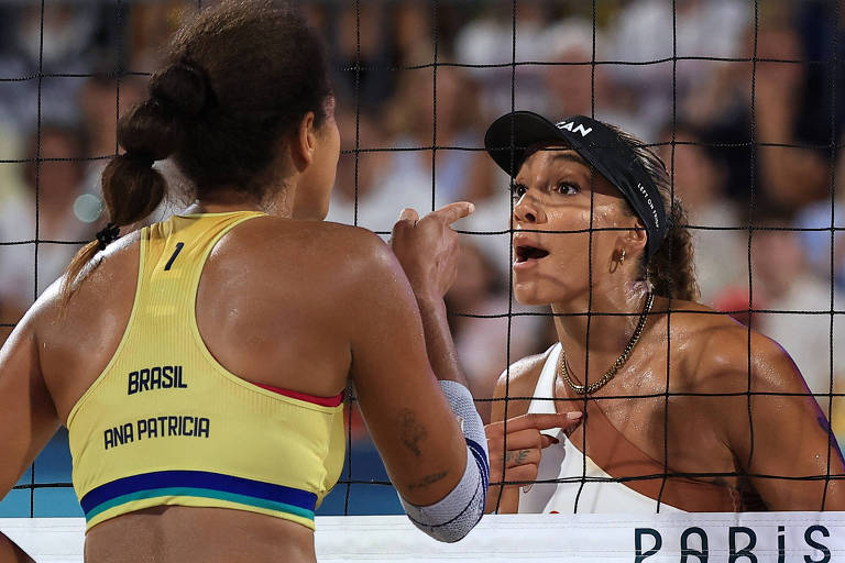 Duas jogadoras de vôlei de praia estão se confrontando na rede. A jogadora à esquerda, com a camisa amarela, tem o nome 'ANA PATRICIA' e a bandeira do Brasil em sua roupa. Ela está de costas, gesticulando com a mão. A jogadora à direita, com uma blusa branca e boné preto, está olhando intensamente para a outra, com a boca aberta, como se estivesse falando. O fundo mostra uma multidão assistindo ao jogo.