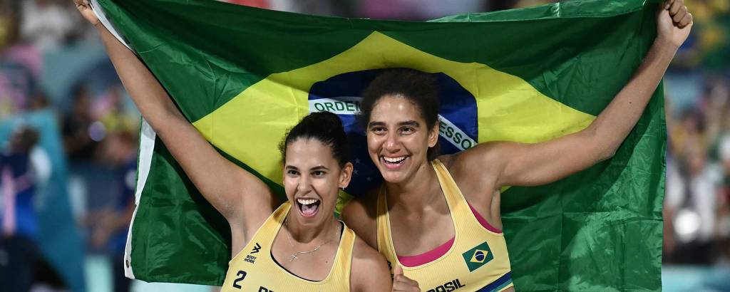 Duas mulheres sorridentes estão segurando a bandeira do Brasil. Elas vestem tops amarelos com detalhes verdes e estão em um ambiente festivo, possivelmente em um evento esportivo. O fundo mostra uma multidão, sugerindo uma atmosfera de celebração.