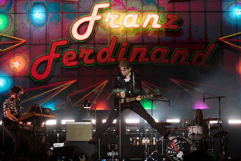 no centro da foto, um homem toca guitarra. há um painel de luzes escrito "Franz Ferdinand"