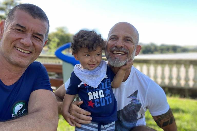 Dois homens sorriem junto de um menino, que aparenta ter cerca de dois anos. Os três estão em um local aberto, com grama verde e céu azul. O homem à esquerda usa uma camiseta de manga curta azul e o homem da direita usa uma camiseta branca. No centro, o menino usa um babeiro azul claro e uma camiseta azul escura. 