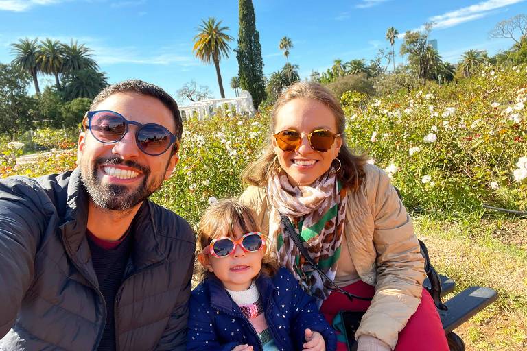 Fotografia mostra uma família sentada em um ambiente de natureza, com céu azul e plantas verdes ao redor. O homem tem pele clara, barba e cabelo escuro, usa uma jaqueta e um óculos de sol. Ao lado dele está uma mulher de cabelos claros, óculos de sol e uma jaqueta bege. Entre eles está uma menina, que aparenta ter dois anos, de óculos de sol cor de rosa e jaqueta azul. Os três estão sorrindo.