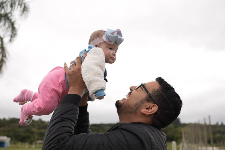 Um homem de cabelo escuro e óculos segura um bebê acima do rosto. Ele está olhando para a criança e sorrindo. A menina usa uma calça rosa e um laço na cabeça.