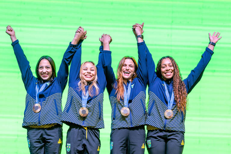 Quatro atletas estão em pé, com os braços levantados em celebração. Todas estão vestindo jaquetas azuis e calças escuras, e cada uma delas exibe uma medalha pendurada no pescoço. O fundo é de uma cor verde vibrante.
