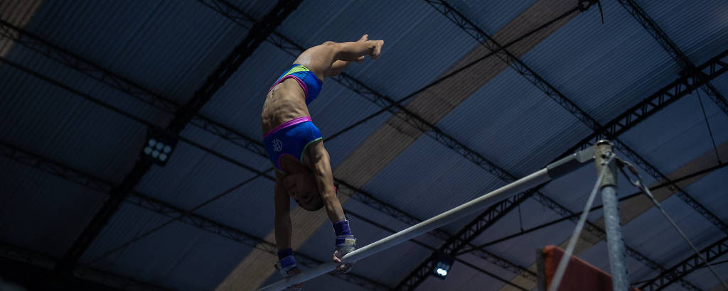 Um atleta está realizando um movimento na barra fixa, com o corpo em posição invertida. Ele usa um traje de ginástica azul e está em um ambiente interno com um teto alto. A iluminação é suave, destacando a figura do atleta em ação.
