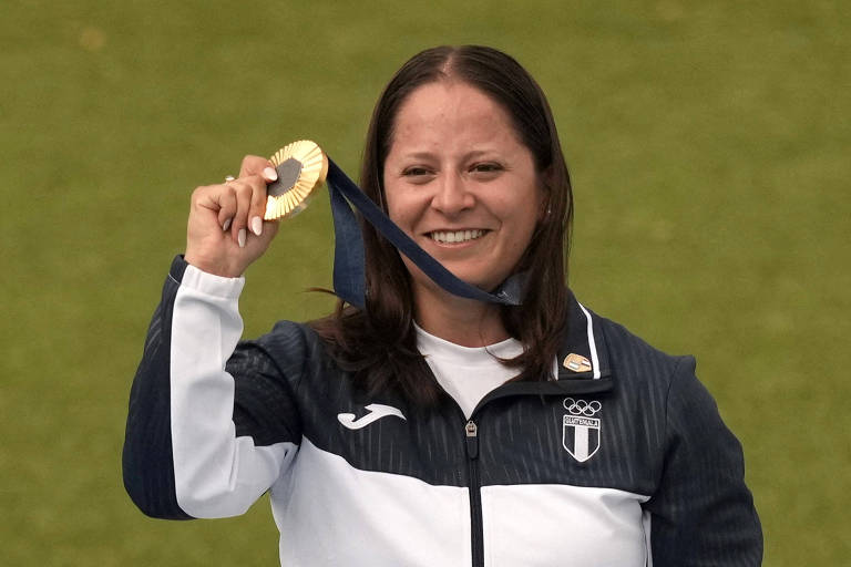 Uma atleta está sorrindo enquanto segura uma medalha de ouro em sua mão. Ela está vestindo uma jaqueta esportiva de cores escuras e claras, com um logotipo visível. O fundo é um campo verde.