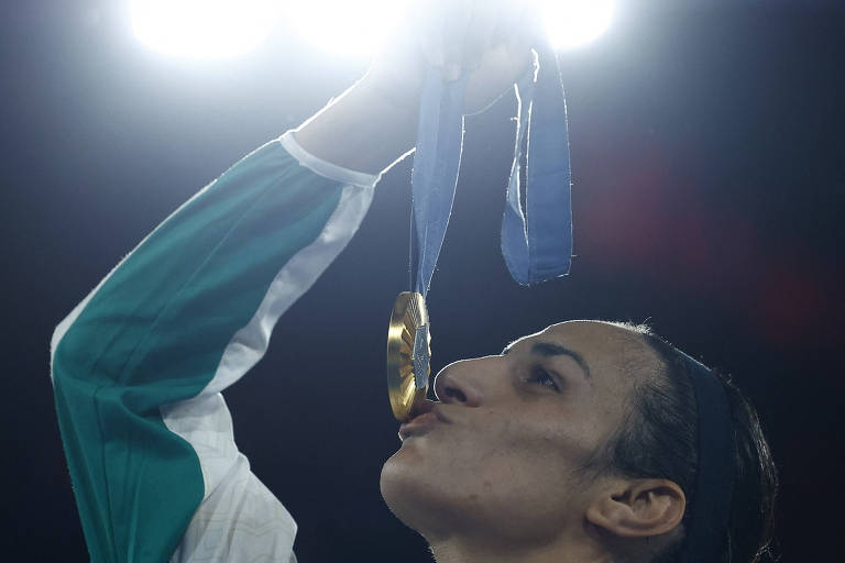 A imagem mostra uma atleta segurando uma medalha de ouro e beijando-a. Ela está vestindo uma jaqueta branca com detalhes em verde. A iluminação intensa do fundo destaca seu rosto e a medalha. O ambiente parece ser um evento esportivo.
