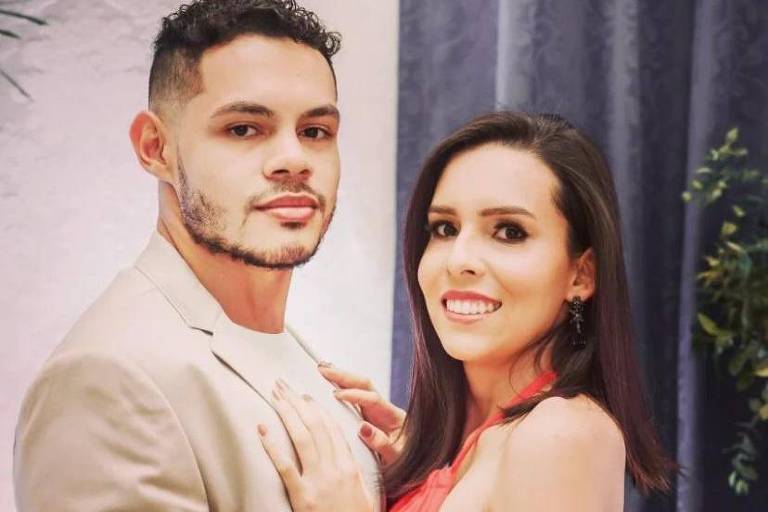 Casal está abraçado e sorri para foto. Ela veste um vestido rosa e ele está de terno.