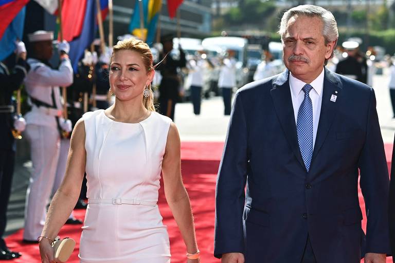 A imagem mostra um evento oficial com um homem e uma mulher caminhando em um tapete vermelho. A mulher está vestindo um vestido branco e segurando uma bolsa, enquanto o homem está usando um terno escuro com uma gravata clara. Ao fundo, há bandeiras de diferentes países e uma formação de guardas.