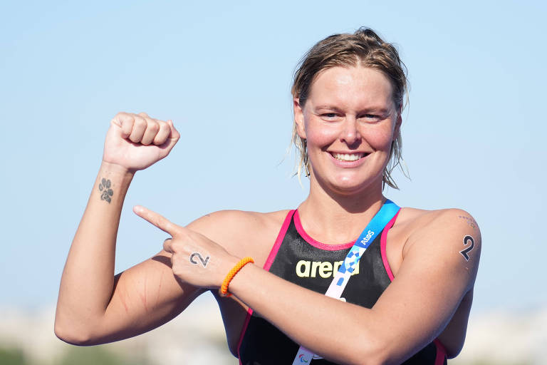 Uma atleta sorri e aponta para o seu braço, onde há um tatuagem visível, com gomos da pata de um cachorro. Ela está usando um traje de competição e uma medalha ao redor do pescoço. O fundo é claro, sugerindo um dia ensolarado.
