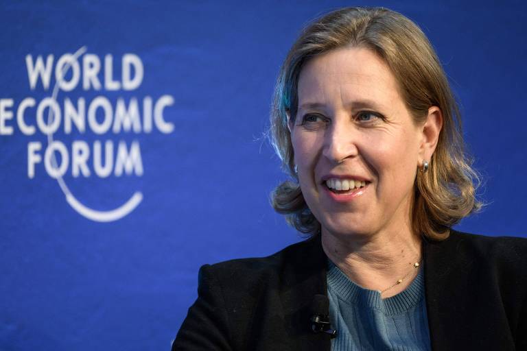 A imagem mostra Susan Wojcicki, uma mulher com cabelo loiro e preso, vestindo uma blusa azul e um blazer preto. Ela está sorrindo e sentada em um evento, com um fundo azul onde está escrito 'Fórum Econômico Mundial' em letras brancas.