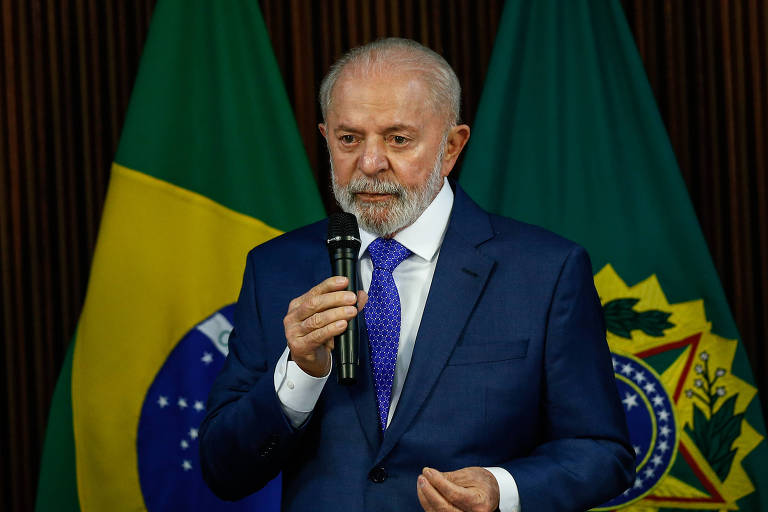 Homem branco vestido de terno gesticula com microfone na mão. Atrás dele há duas bandeiras do Brasil