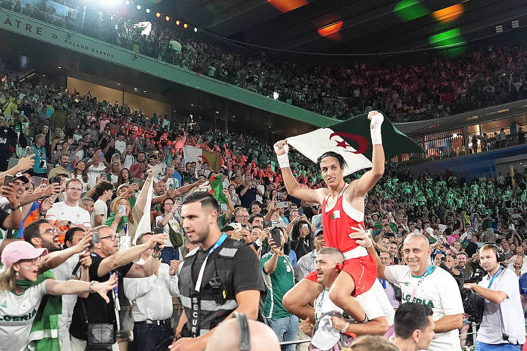 A imagem mostra uma multidão vibrante em um evento esportivo em Roland Garros. No centro, um atleta em um uniforme vermelho é carregado por um homem, segurando uma bandeira da Argélia. A plateia ao redor está animada, com pessoas levantando os braços e segurando celulares para registrar o momento. O ambiente é festivo, com várias bandeiras e torcedores visivelmente empolgados