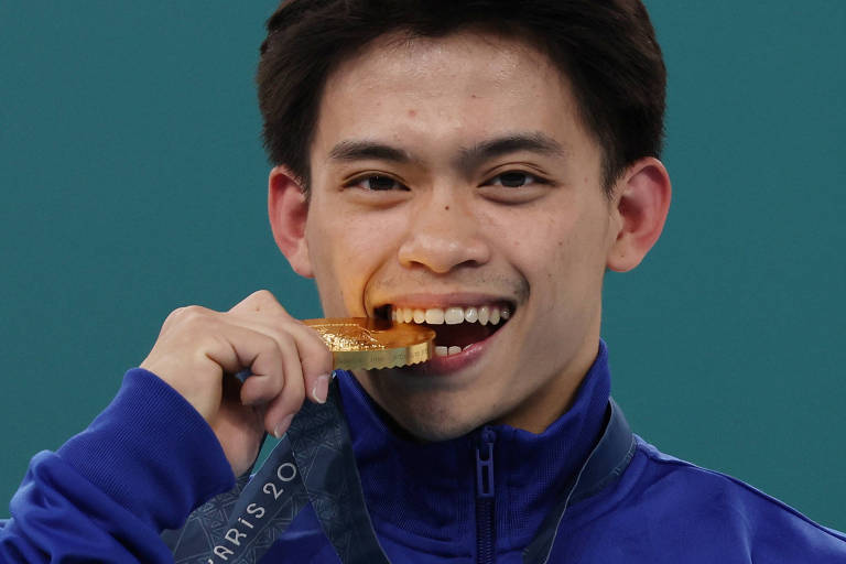 Um atleta sorridente está segurando uma medalha de ouro na boca, enquanto exibe um sorriso. Ele usa uma jaqueta azul e tem cabelo escuro. O fundo é de uma cor verde sólida.