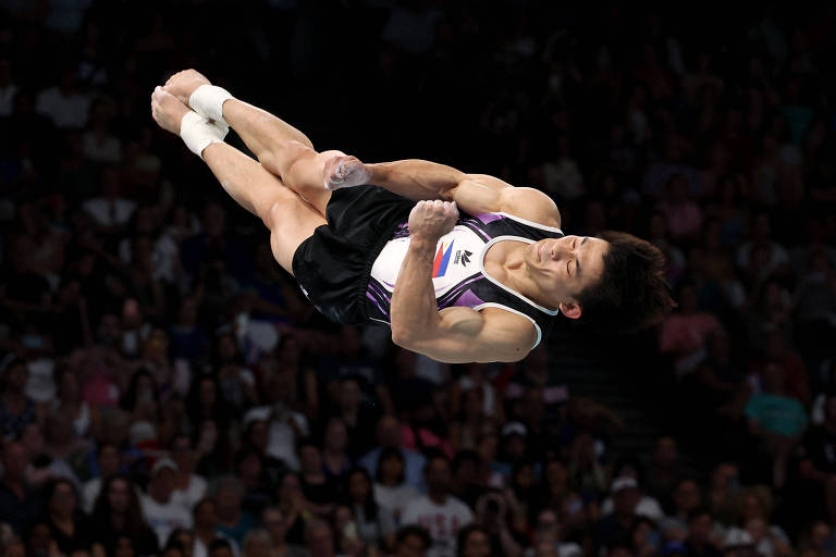 Um ginasta está realizando um salto acrobático no ar, com o corpo em posição horizontal e as pernas estendidas. Ele usa um traje de competição com detalhes em roxo e uma bandeira nas costas. O fundo mostra uma plateia cheia, com pessoas assistindo ao evento.