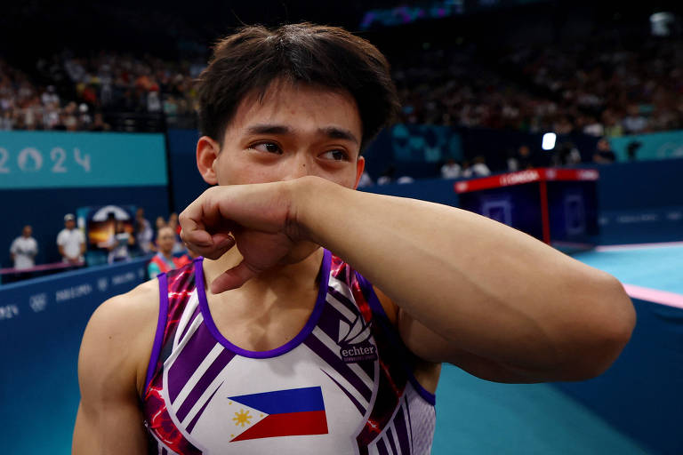 Um atleta de ginástica, vestindo um traje com as cores da bandeira das Filipinas, está se limpando com o braço enquanto expressa emoção após uma apresentação. O fundo mostra uma multidão e um ambiente de competição, com equipamentos de ginástica visíveis.