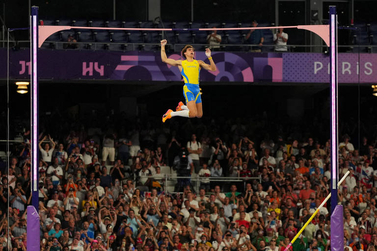 Um atleta está no ar, realizando um salto com vara. Ele usa um uniforme amarelo e azul, representando a Suécia. O fundo mostra uma multidão animada, com pessoas aplaudindo e tirando fotos. O atleta está prestes a passar sobre a barra, que está posicionada em suportes altos. O ambiente é de uma competição esportiva, com iluminação intensa e um grande número de espectadores.