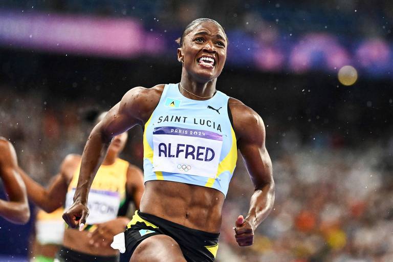 A imagem mostra uma atleta correndo em uma competição, com um olhar determinado. Ela veste uma camiseta azul clara com detalhes em amarelo e preto, representando Saint Lucia, e tem o nome 'ALFRED' visível em seu uniforme. O fundo é desfocado, sugerindo um ambiente de competição intensa, e há gotas de suor visíveis em seu corpo.