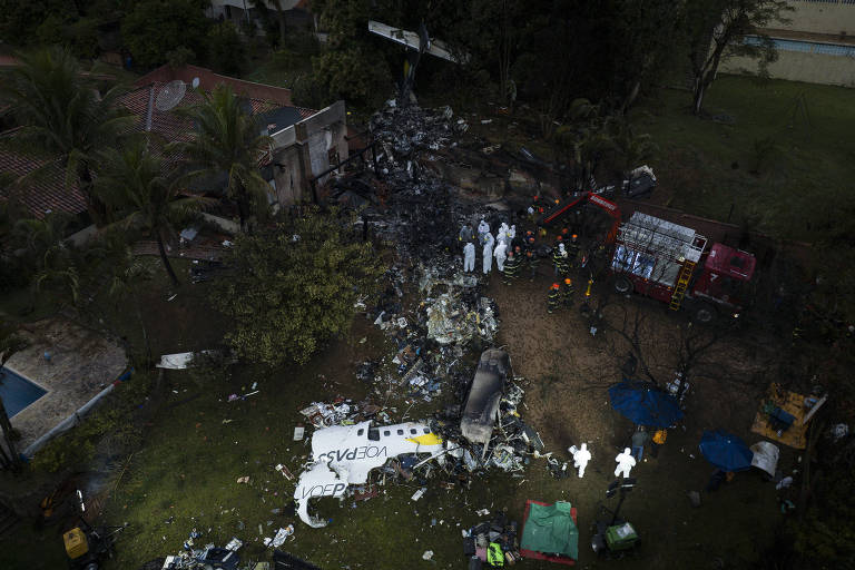 Imagem aérea mostra uma área com muitos destroços queimados e algumas pessoas com roupas de proteção branca caminhando entre eles; na parte baixa é possível ver uma parte maior do avião