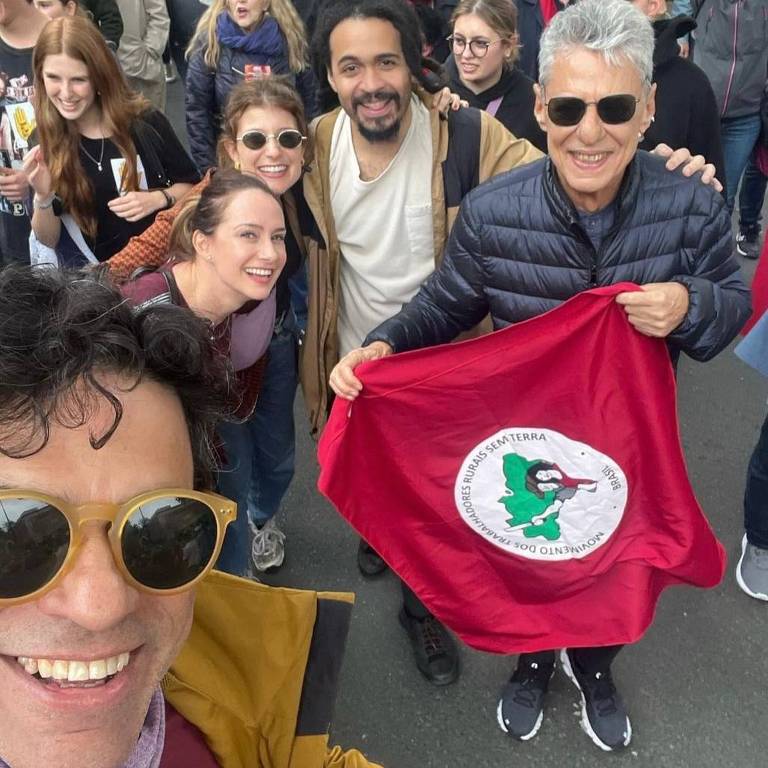 A imagem mostra um grupo de pessoas sorrindo em uma manifestação. No primeiro plano, um homem com óculos escuros e uma jaqueta amarela está tirando uma selfie. Ao seu lado, há três mulheres e um homem segurando uma bandeira vermelha com um símbolo do MST (Movimento dos Trabalhadores Rurais sem Terra). Ao fundo, várias outras pessoas estão visíveis, algumas acenando e outras sorrindo. O ambiente parece ser ao ar livre, com um clima de celebração