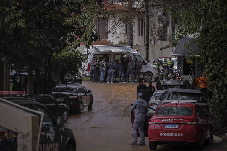 A imagem mostra uma rua com vários veículos estacionados, incluindo carros de polícia. Há um grupo de pessoas, possivelmente policiais, reunidos em torno de uma van branca. O ambiente parece ser urbano, com árvores e edifícios ao fundo. O clima parece nublado ou chuvoso.