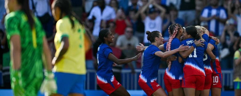 A imagem mostra um grupo de jogadoras de futebol comemorando um gol. Elas estão vestidas com uniformes azuis e vermelhos, cercadas por torcedores ao fundo. À esquerda, uma jogadora da equipe adversária, vestindo um uniforme verde, observa a celebração. O ambiente é de festa e emoção, com a torcida ao fundo.