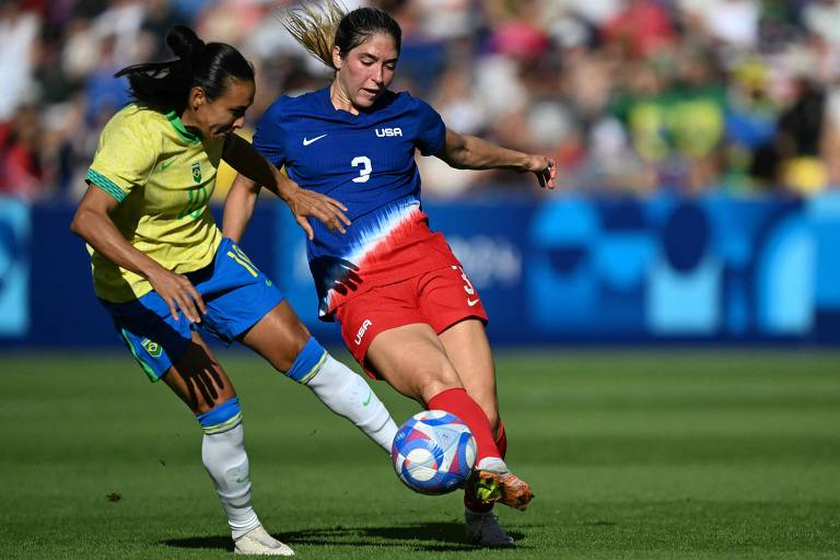 A imagem mostra duas jogadoras de futebol em um momento de disputa pela bola. A jogadora à esquerda está vestindo um uniforme amarelo e azul, enquanto a jogadora à direita usa um uniforme vermelho e azul. Ambas estão em um campo de futebol, com uma multidão ao fundo.
