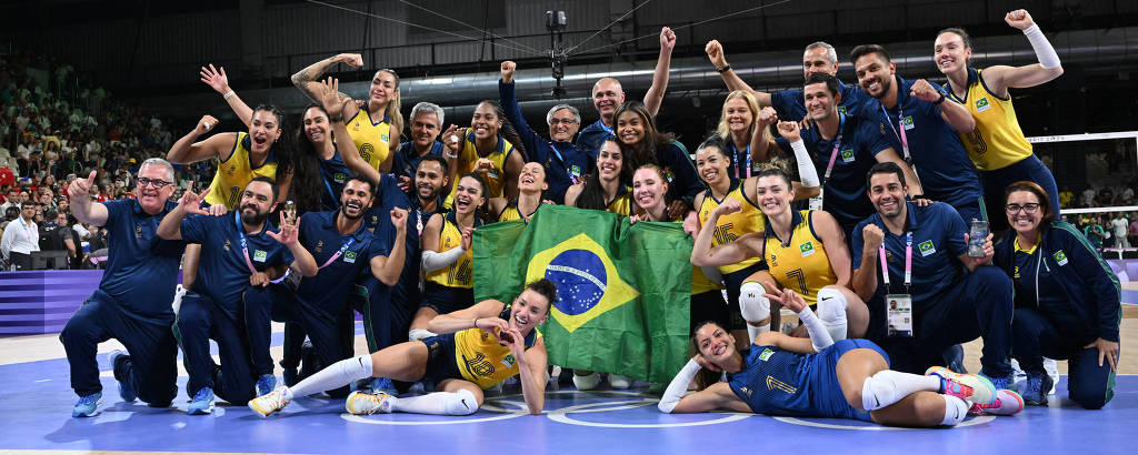 A imagem mostra a equipe de vôlei feminino do Brasil comemorando em uma quadra. As jogadoras estão vestindo uniformes amarelos e azuis, algumas segurando uma bandeira do Brasil. O ambiente é festivo, com sorrisos e gestos de celebração. Ao fundo, há uma tela exibindo a bandeira do Brasil.