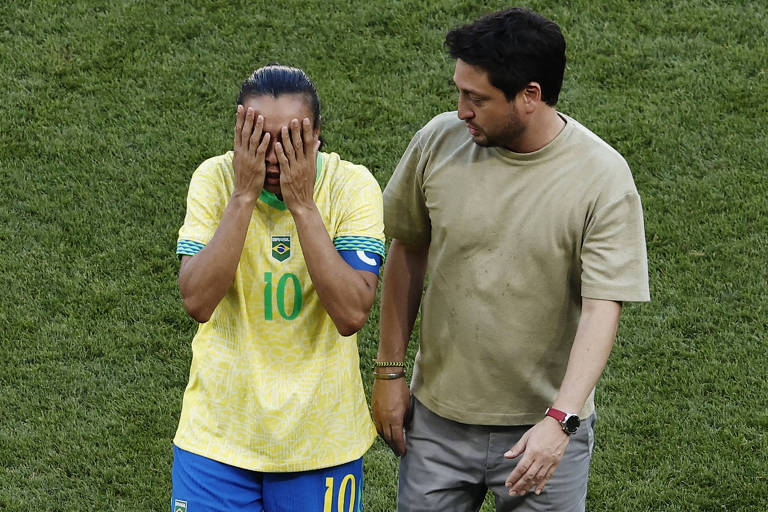 A imagem mostra uma jogadora de futebol vestindo uma camisa amarela com o número 10, cobrindo o rosto com as mãos, aparentando estar emocionada. Ao seu lado, um homem vestido com uma camiseta bege observa e parece estar oferecendo apoio. O fundo é um campo de futebol com grama verde.
