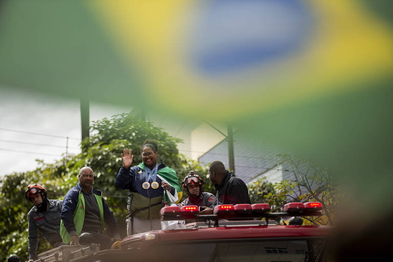 A imagem mostra um grupo de pessoas em um carro, acenando e sorrindo. Eles estão cercados por uma vegetação verde e há uma bandeira do Brasil em primeiro plano, desfocada. Os indivíduos parecem estar celebrando, possivelmente em um evento público ou desfile.
