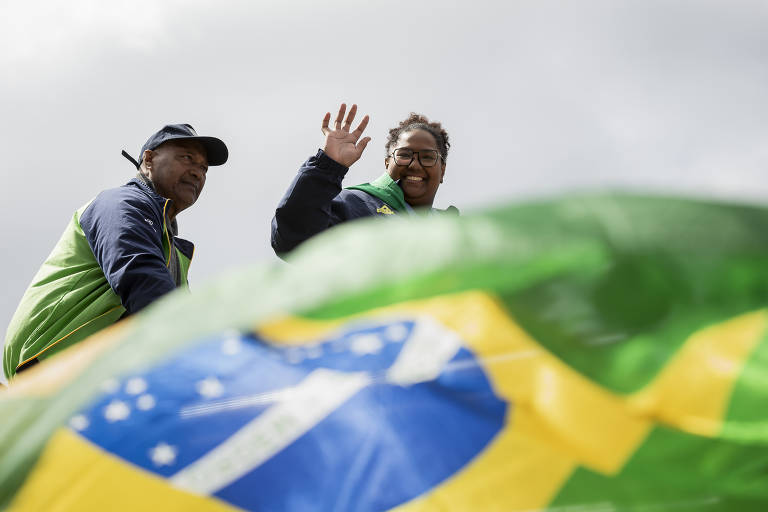 Duas pessoas estão acenando, uma delas com um sorriso. Elas estão vestidas com roupas em tons de verde e azul. Ao fundo, uma bandeira do Brasil é visível, com as cores verde, amarelo, azul e branco. O céu está nublado.