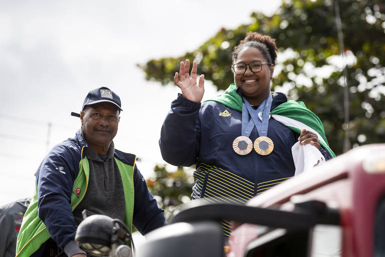 A imagem mostra uma mulher sorridente acenando com a mão, usando um moletom escuro e com duas medalhas penduradas no pescoço. Ao lado dela, um homem com um boné e uma jaqueta azul e verde está sentado em um veículo. Ao fundo, há árvores e um céu nublado.