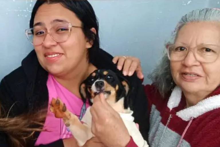 Duas mulheres, sendo uma idosa, seguram nos braços um cão de pelo branco e preto