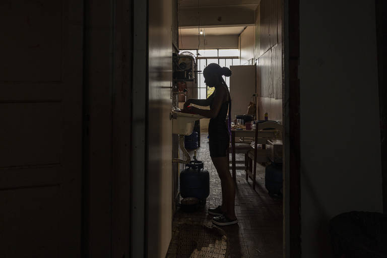 Foto em contra-luz mostra jovem lavando a louça em casa escura, com janela ao fundo