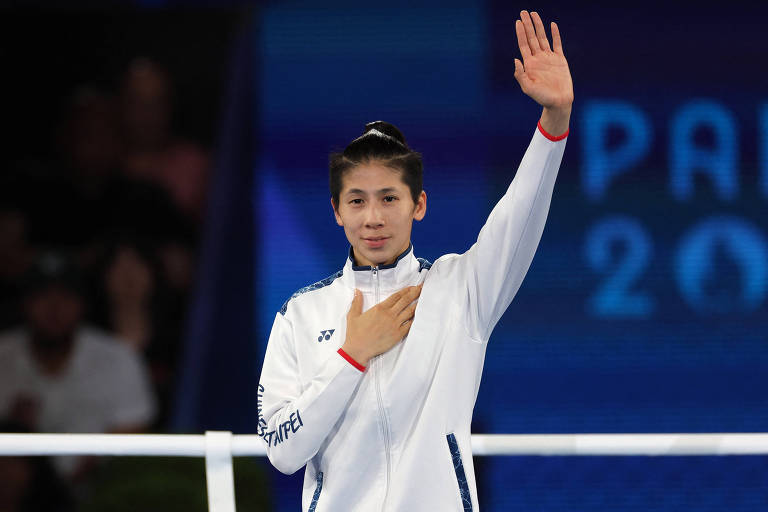 Uma atleta está em pé, vestindo uma jaqueta branca com detalhes em azul e um símbolo de marca. Ela está acenando com a mão direita levantada e a mão esquerda sobre o coração, em um gesto de saudação. O fundo é desfocado, mas há uma tela que exibe 'PARIS 2024'.
