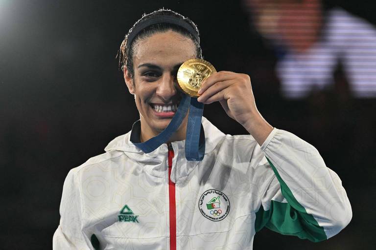 Uma atleta sorri enquanto segura uma medalha de ouro em frente ao rosto, com um fundo desfocado. Ela está vestindo uma jaqueta branca com detalhes em verde e vermelho, representando a bandeira de seu país. A medalha tem um laço azul e apresenta um design detalhado.
