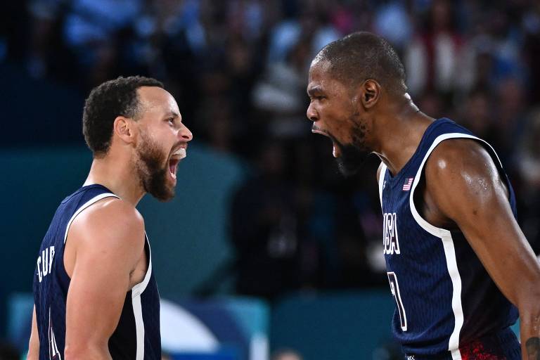 Dois jogadores de basquete estão se enfrentando em uma partida, ambos com expressões intensas. Um jogador à esquerda está gritando, enquanto o jogador à direita também parece estar expressando emoção. Eles vestem uniformes de basquete escuros e estão em um ambiente de jogo com uma multidão ao fundo.
