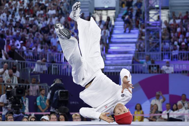 Um dançarino de breaking está realizando uma manobra acrobática em uma competição, com o corpo em posição invertida. Ele veste um traje branco e um gorro vermelho. Ao fundo, uma grande plateia observa, com câmeras de filmagem visíveis.
